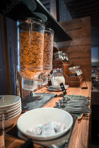 Wooden counter holds cereal dispensers, bowls, plates, cutlery, milk carafe, and jam jars. Labeled signs and warm light suggest a calm early morning alpine chalet ritual.