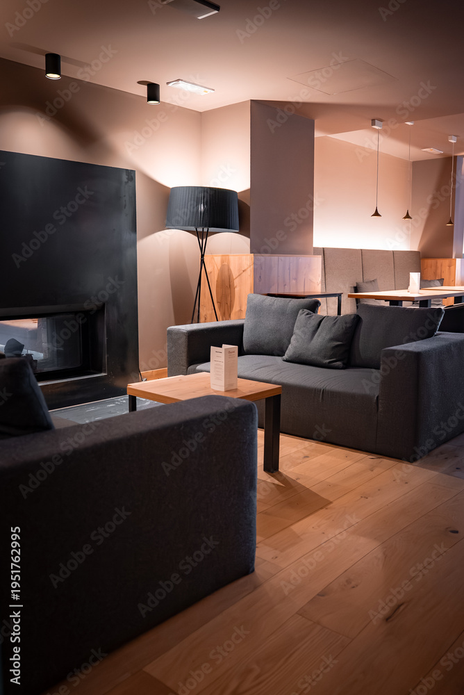 Naklejka premium Charcoal gray sofas circle a low wooden table by a matte black fireplace in San Domenico, Italian Alps. Warm pendant and tripod lamp lighting set an evening mood.