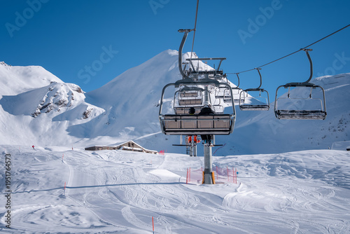 Bild auf Leinwand San Domenico ski resort in the Italian Alps shows a chairlift ascending over groomed pistes, two skiers on the central chair, orange piste markers, lodge, and clear blue skies