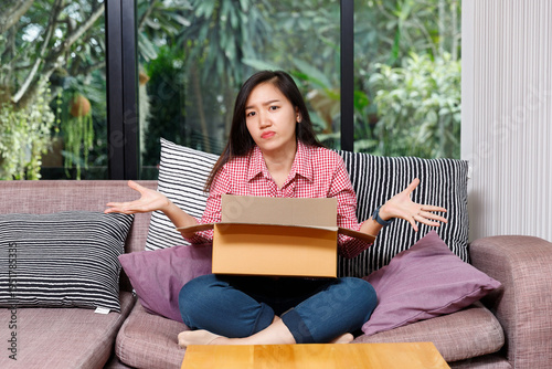 Confused young Asian woman sitting on sofa with an open parcel box and shrugging shoulders. Concept of missing item, wrong order, or online shopping mistake.