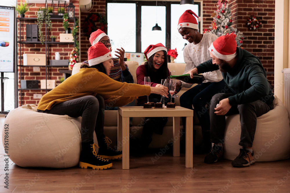 Obraz premium Cheerful people pouring wine in glasses at christmas party in office decorated with festive ornaments. Colleagues celebrating xmas holiday at festivity with alcohol drink and decorations.