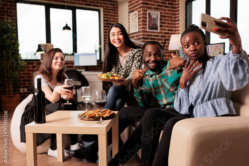 Distinct ethnicities happy people eating snacks, drinking wine, posing for smartphone selfie. African american woman taking selfie with friends at apartment birthday party.