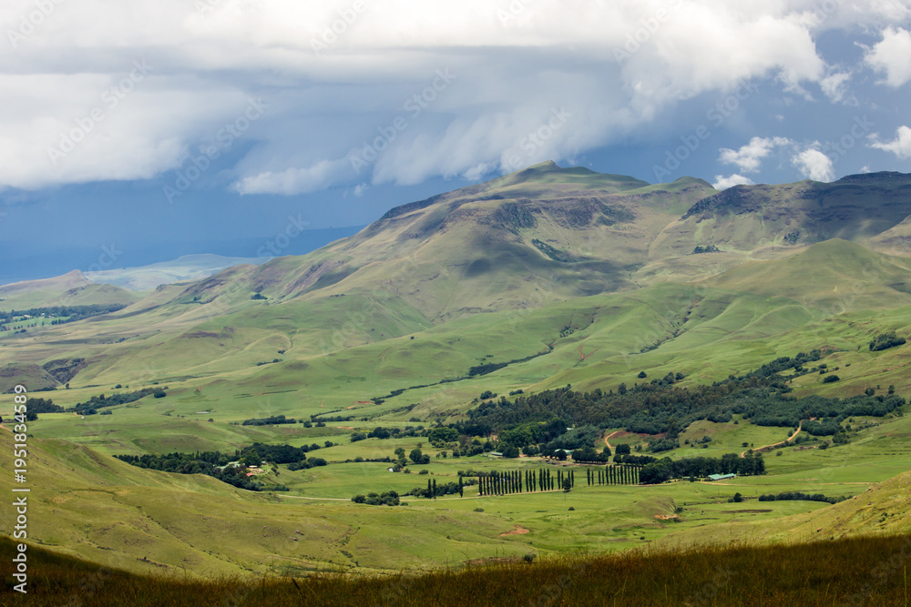 Obraz premium View over bushman’s neck, nestled in a secluded sunlit green valley in the Drakensberg mountains of South Africa, with ominous Strom clouds gathering in a blue-grey sky in the background.