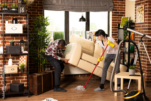 Photos Caucasian woman wearing yellow rubber gloves mopping under sofa as african american husband gives assistance
