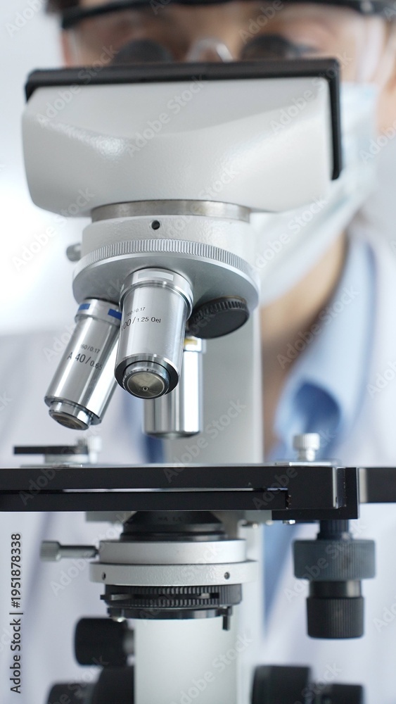 Fototapeta premium Scientist wearing lab coat and protective glasses using a modern microscope in a laboratory setting, conducting research and experiments in a scientific environment. Health care and science concept