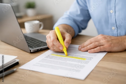 Close-up of hands using a yellow highlighter to mark passages on a printed document on a wooden desk beside an open laptop and coffee cup, showing focused office review and editing