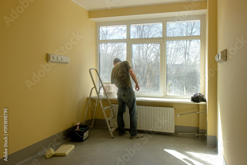 Obraz na płótnie A worker performs repair work near a window from behind in a renovated room with natural sunlight