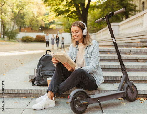 A young adult Caucasian woman is seated on outdoor steps, engrossed in a book while wearing headphones. An electric scooter is parked nearby as she focuses on studying or relaxing during her break.