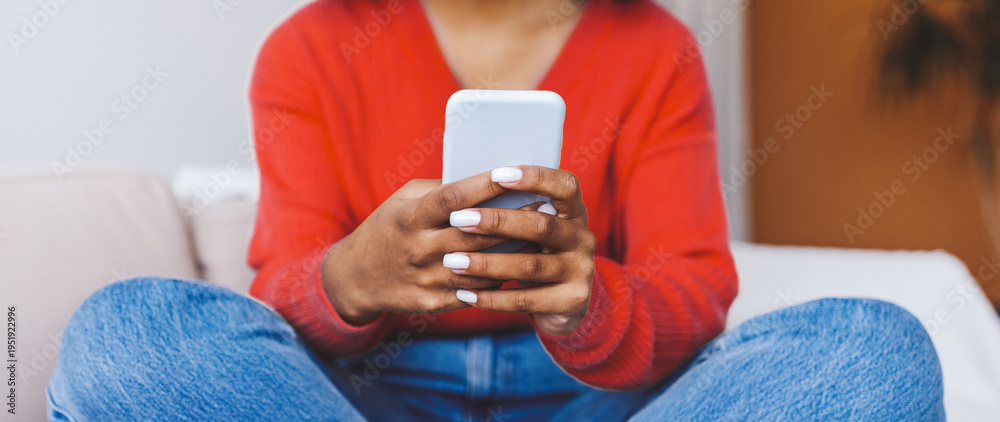 Fototapeta premium A woman sits with her legs crossed on a sofa, focused on her smartphone. She wears a red sweater and jeans. The background shows a cozy living room with plants.