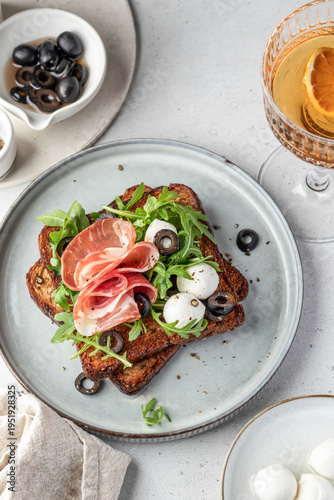 Roasted toasts with prosciutto ham, mozzarella balls, arugula and sliced olives on a plate close up on white table background top view. Delicious breakfast