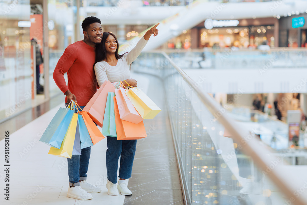 Obraz premium A man and woman are standing in a shopping mall. They hold colorful shopping bags. The man points toward something in the distance while the woman smiles.