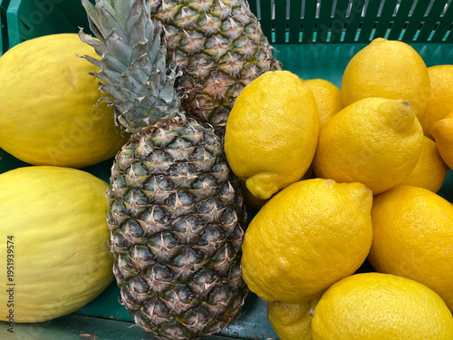 Close‑up of fresh pineapples, yellow melons, and lemons displayed in a green market crate, showcasing vibrant colors and a mix of tropical and citrus fruits.