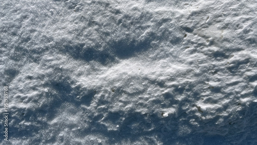 Close‑up of uneven, sunlit snow with fine crystal texture and natural ripples, creating an abstract winter surface with soft shadows and sparkling highlights.