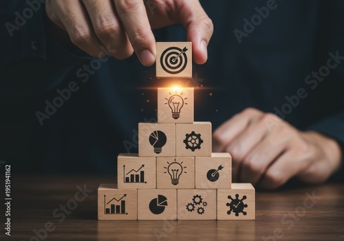 Businessman hand stacking wooden blocks into a pyramid with business icons and placing the target icon on top for strategy planning and goal achievement concept