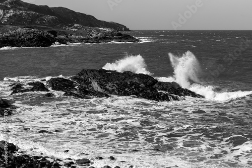 Monotone view of powerful sea spray and churning ocean water hitting a rocky shoreline