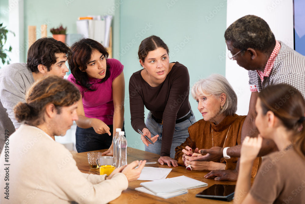 Obraz premium Group of people of different ages are sitting at a table and talking to each other, they are having a discussion and looking at documents. Office employees discuss the launch of the project