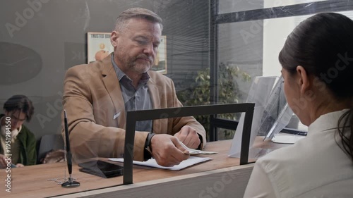 Middle-aged male customer handing cash money to bank teller across glass counter in modern bank office