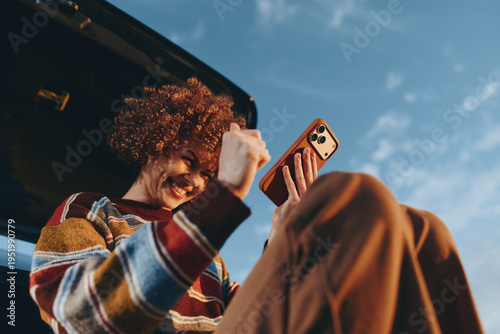 Fotografía Smiling woman with curly hair wearing a rainbow sweater sitting in a car trunk holding a smartphone, enjoying a sunny day with clear blue sky background, lifestyle and happiness concept
