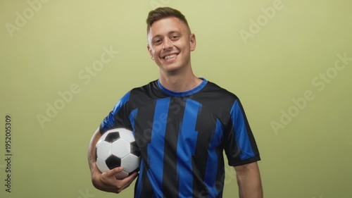 Young blond man holds soccer ball under arm, smiling and pointing with free hand while facing camera in studio; confidence.