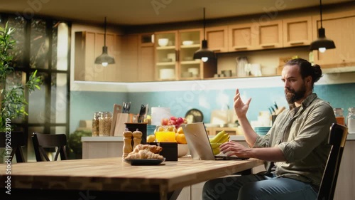 Disoriented man being confused and uncertain with his work at laptop, working from home in the kitchen. Male freelancer unsure frowning at the screen, ambiguous teleworking.