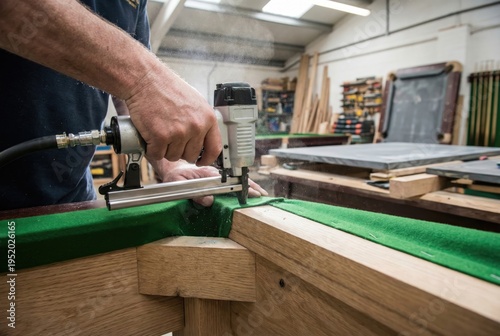Craftsman using a pneumatic staple gun to attach green felt cloth to the wooden frame of a billiard table