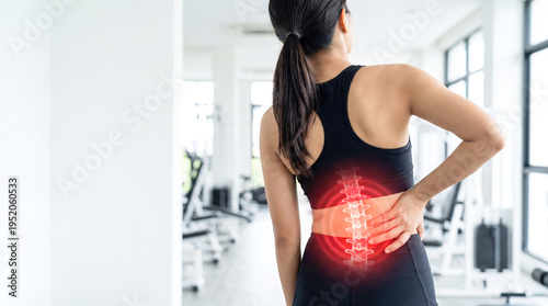 A woman is stretching in a gym and holding her lower back. The room has exercise equipment and large windows that let in light. She looks focused on relief