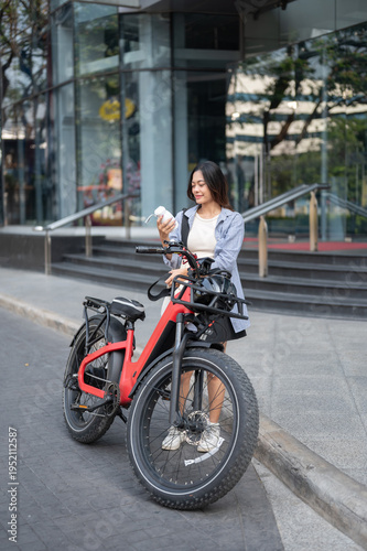 Asian woman holding tumbler standing with fat tire electric bicycle on pavement in front of building