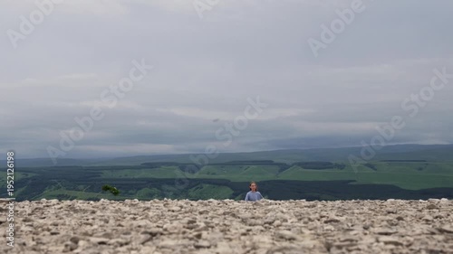 Young woman on rocky plateau gazing out over distant green hills under heavy clouds, wind tousling dress and hair, slow steps across limestone field, atmospheric cinematic mood evoking