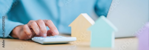 Close up of male hand using a calculator near wooden house models and laptop to analyze mortgage payments, home refinance strategy, and property investment costs