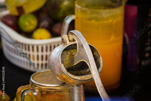 A vintage metal citrus press in close up at a Bangkok street food beverage stall. In the blurred background is a pitcher of fresh orange juice, a basket of fruit and a strainer.