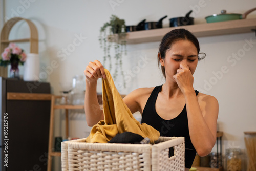 фотографія Woman smelling musty laundry from basket at home, reacting to bad odor from damp