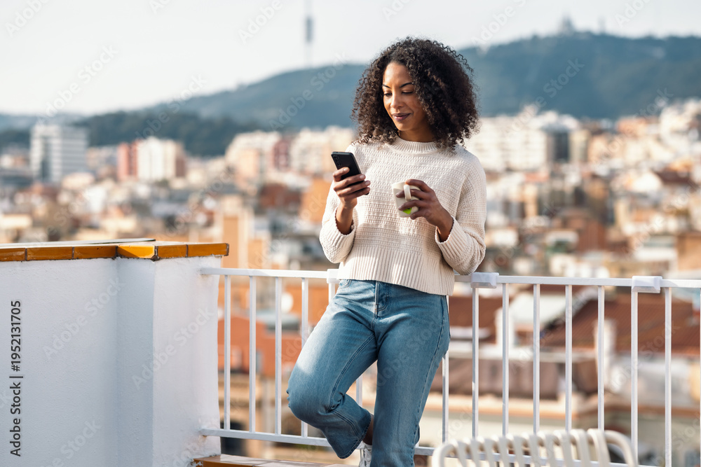 Fototapeta premium Beautiful woman drinking a cup of tea while doing a videocall on her balcony at home