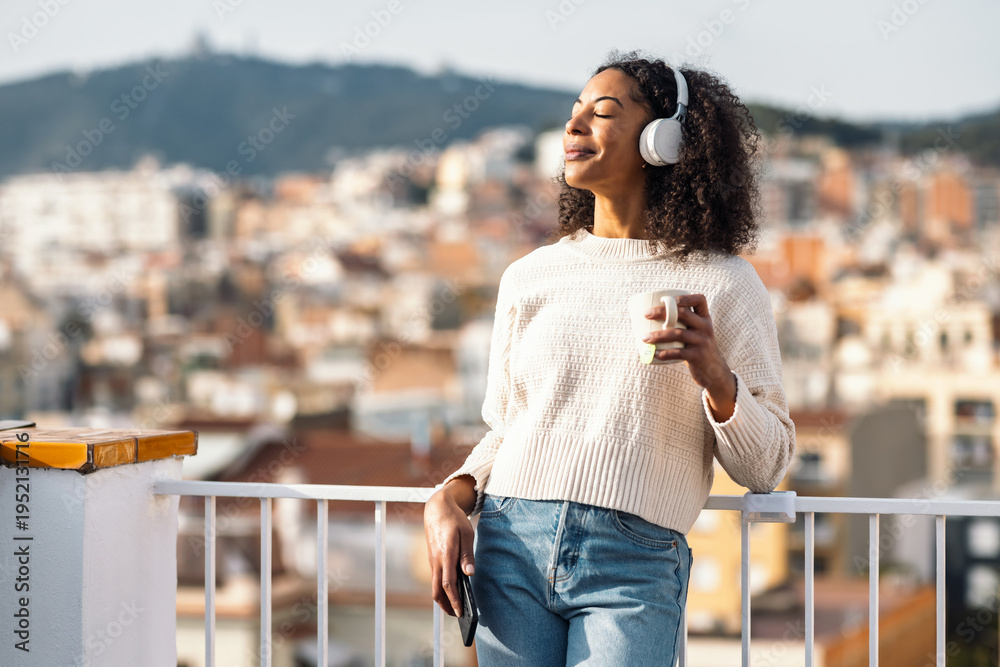 Naklejka premium Relaxing woman drinking a cup of tea while listening to music with headphones on her balcony at home