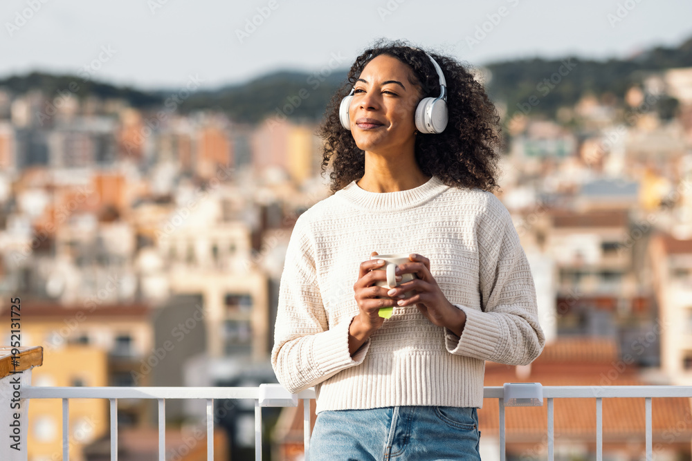 Obraz premium Relaxing woman drinking a cup of tea while listening to music with headphones on her balcony at home