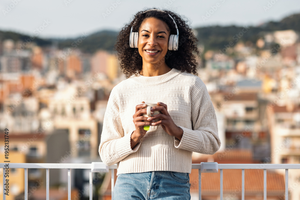 Fototapeta premium Relaxing woman drinking a cup of tea while listening to music with headphones on her balcony at home