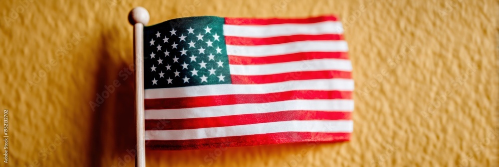 Obraz premium Small American flag on a pole stands against a yellow textured wall background for national themes, government education, and patriotic celebrations with copy space