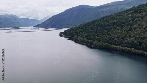 A drone moves sideways above Tingvollfjorden in Norway. It captures the view of the fjord and the mountains in blue haze, the wooded shore, and a road along the bank. Clouds are visible in the sky.
