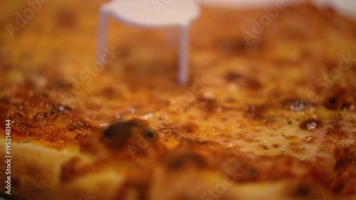 An appetizing close-up view captures a large cheese pizza in a cardboard delivery box. A small plastic pizza saver sits on the golden-brown surface, while a missing slice reveals the white box liner. 
