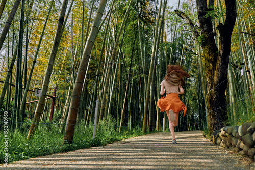 Картината върху платно Bamboo forest path woman running in orange skirt, back view in motion along a gravel trail