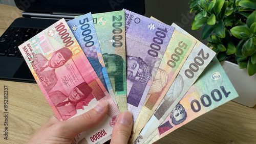 Fotografie A hand holds a fan of Indonesian rupiah banknotes of various denominations against a background of a laptop and a plant