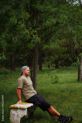 middle-aged muscular, blond, sporty man with a short beard, wearing a baseball cap, black, shorts, green T-shirt, posing in the forest