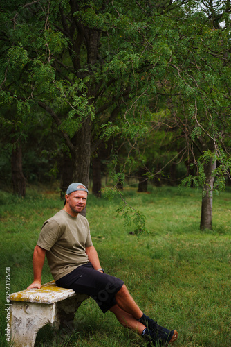 middle-aged muscular, blond, sporty man with a short beard, wearing a baseball cap, black, shorts, green T-shirt, posing in the forest