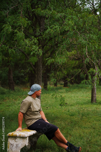 middle-aged muscular, blond, sporty man with a short beard, wearing a baseball cap, black, shorts, green T-shirt, posing in the forest