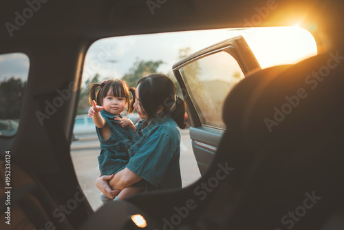 Happy Asian mother holding her young daughter by an open car door during sunset. Concept of family road trip, child safety, and emotional bonding. vacation journey