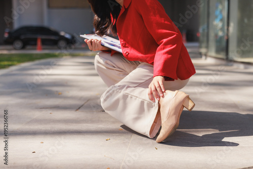 Woman experiencing ankle pain outdoors while holding documents