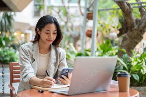 Free Download Asian businesswoman or student looking at phone writing aside laptop sitting at table in garden cafe - GFXLoad