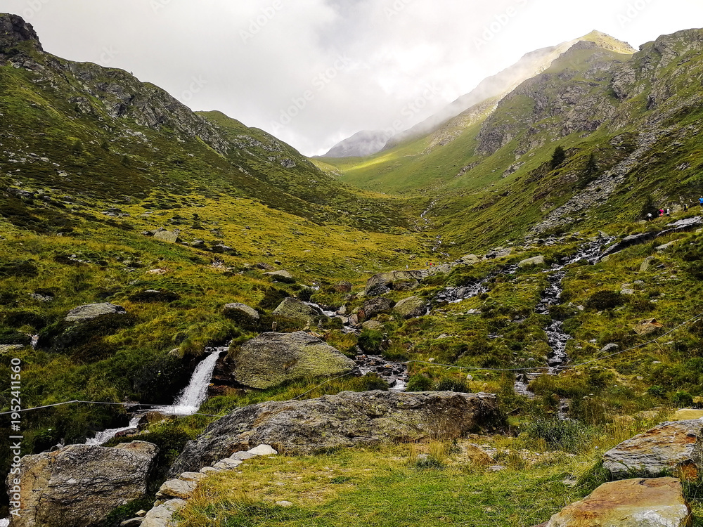 Obraz premium Alpine Valley with Stream and Waterfall Leading to a Mountain Pass Under Dramatic Clouds, Italian Alps Landscape