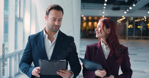 Front view dolly slow motion shot of Latin businesswoman and European businessman walking and discussing business holding tablet and clipboard in modern office corridor