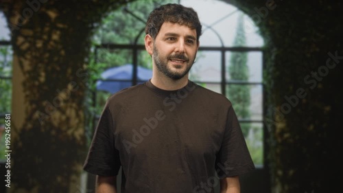 Hispanic man with beard smiling and standing in front of arched window wearing brown t shirt in studio; calm confidence.