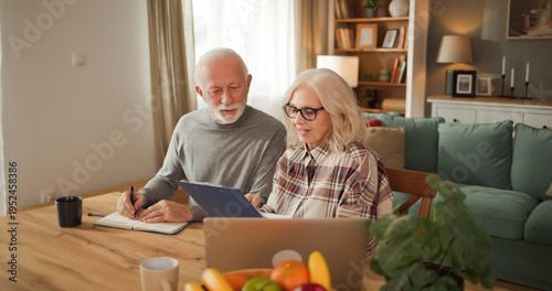 Senior couple works on life insurance planning at home, sitting at a table with a laptop, papers, and fruit, focusing on their financial future together in the living room.